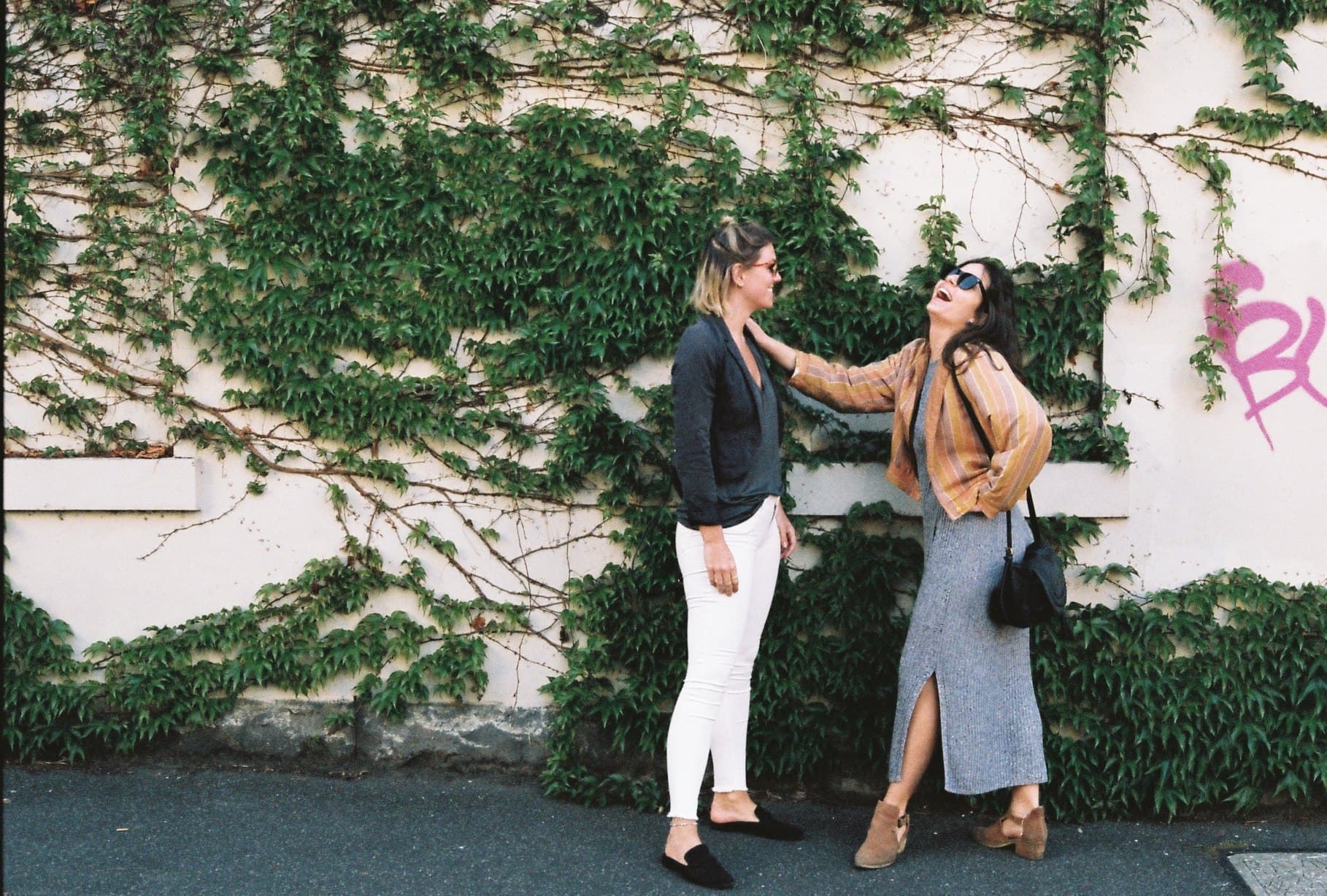 Two people posing beneath trees beside a wall.