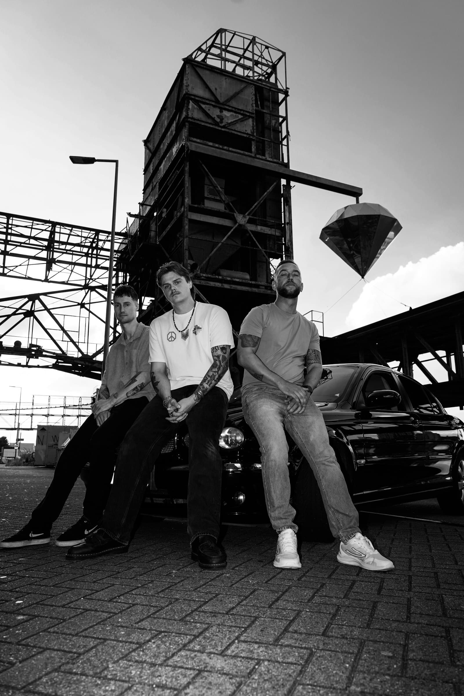 Black-and-white photograph of three Corleone band members leaning against a car beneath industrial steel structures.