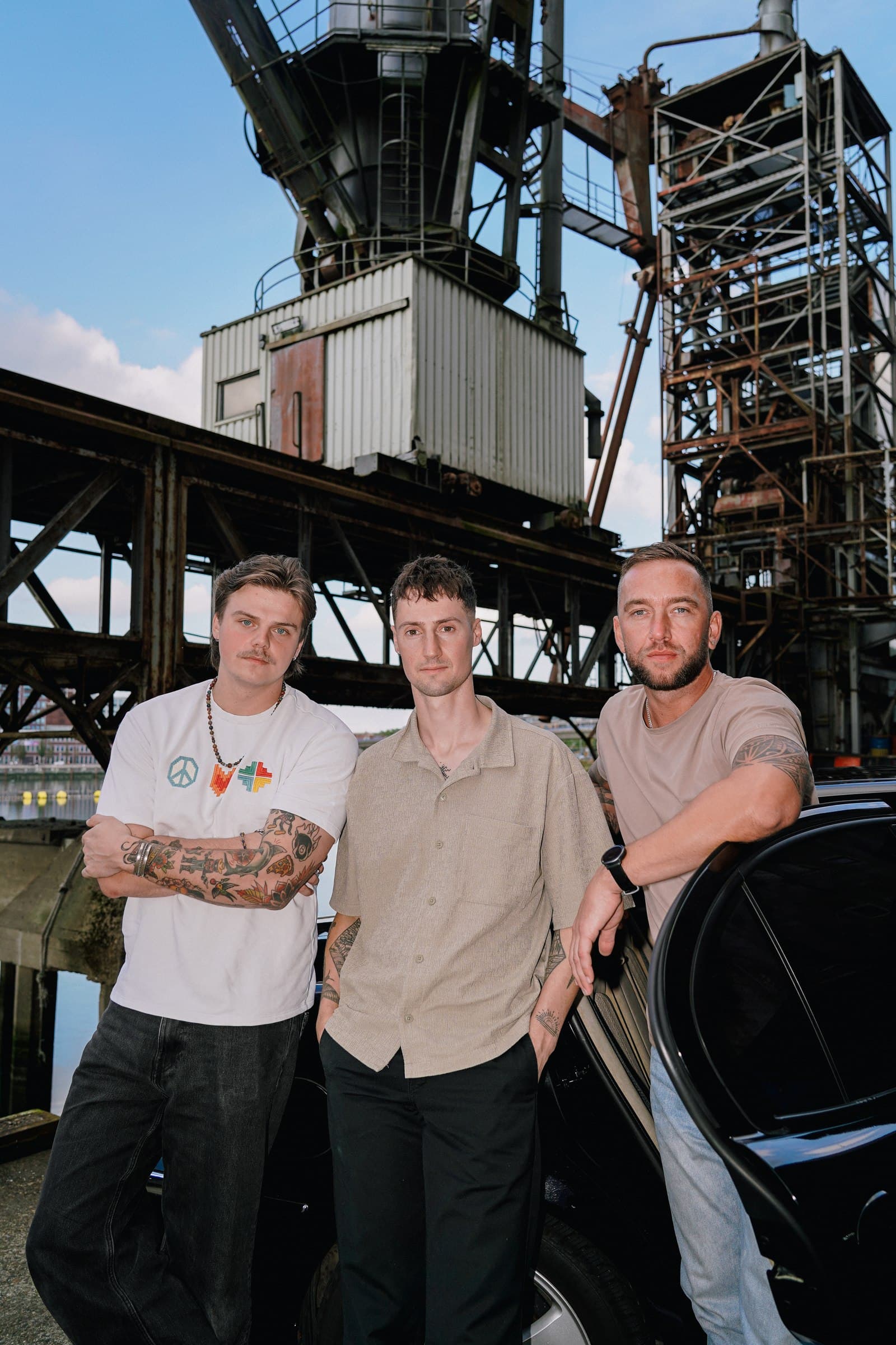 Three Corleone band members standing beside a black car with industrial steelwork behind them.
