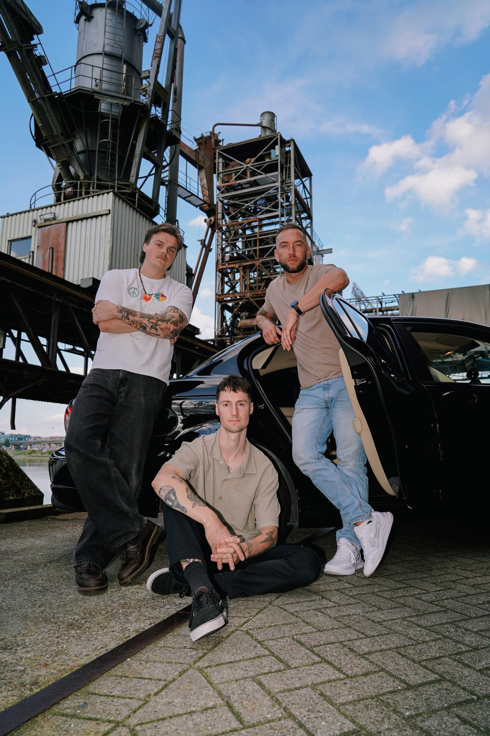 Corleone band members beside a black car beneath industrial steel structures.