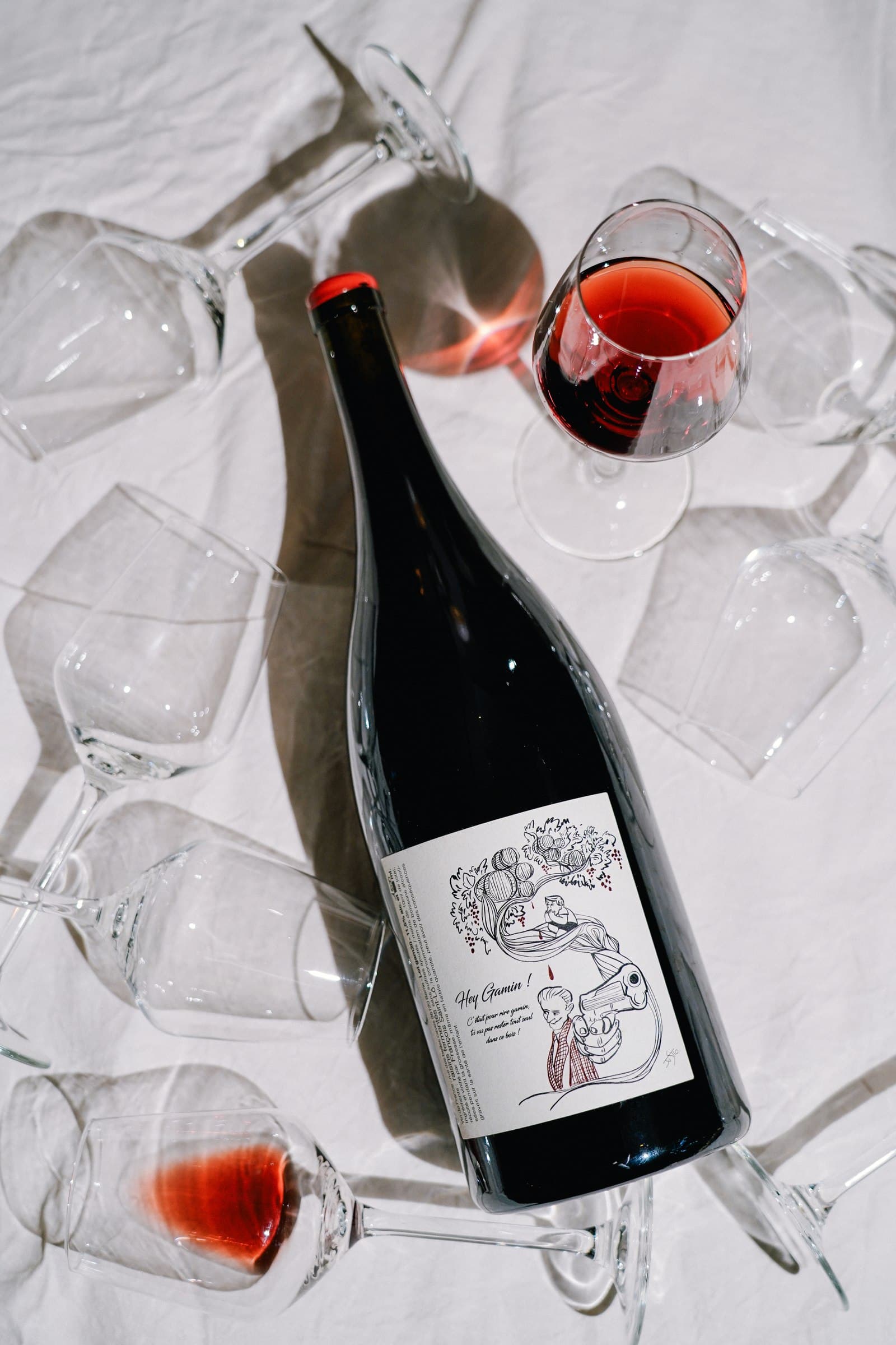 A dark wine bottle and a glass of red wine arranged on a pale textured surface.