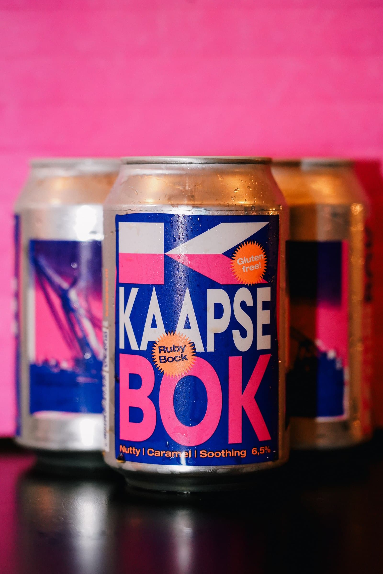 Kaapse Bok beer cans arranged against a bright pink background.