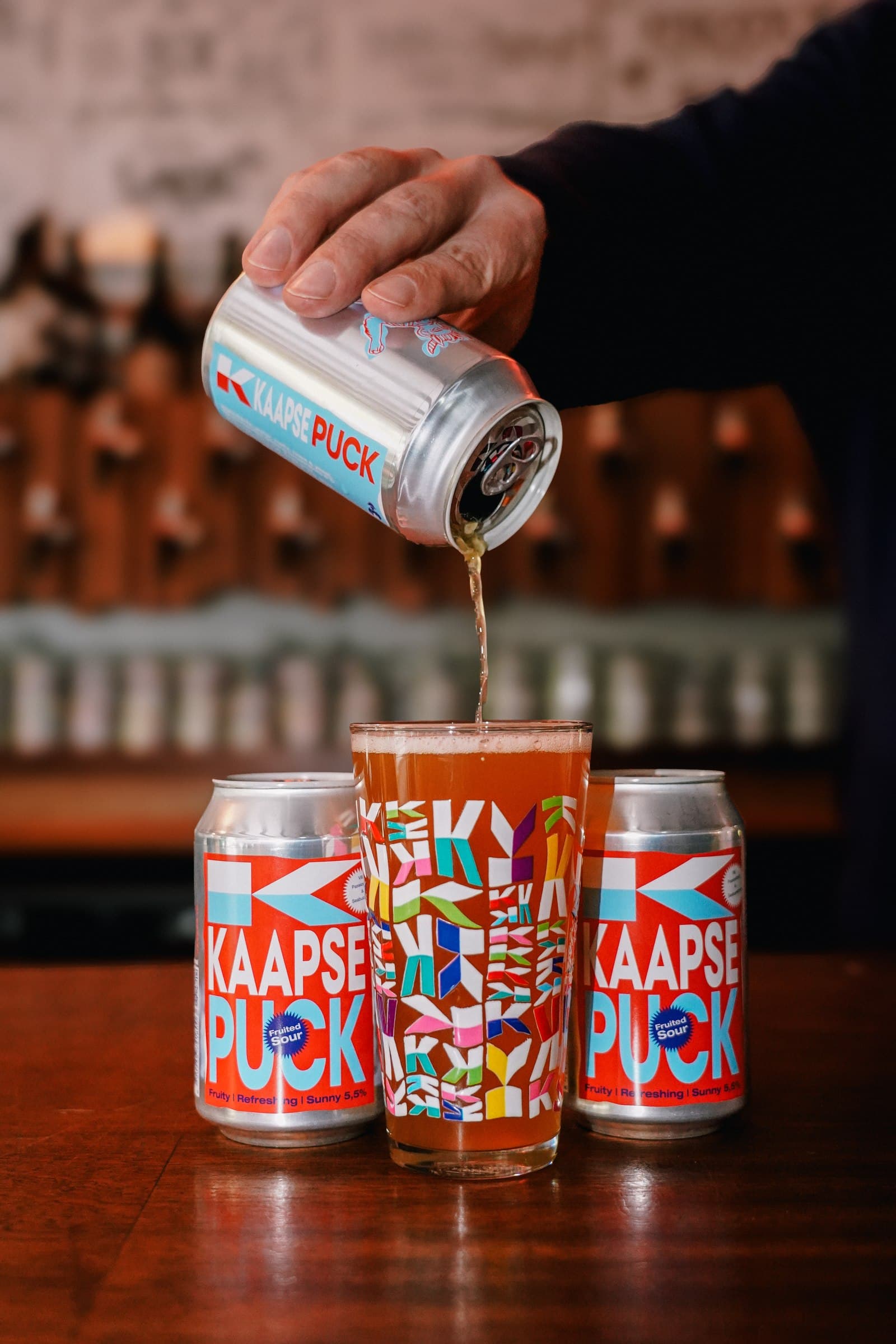 A hand pouring a drink above illustrated beer cans on a bar counter.