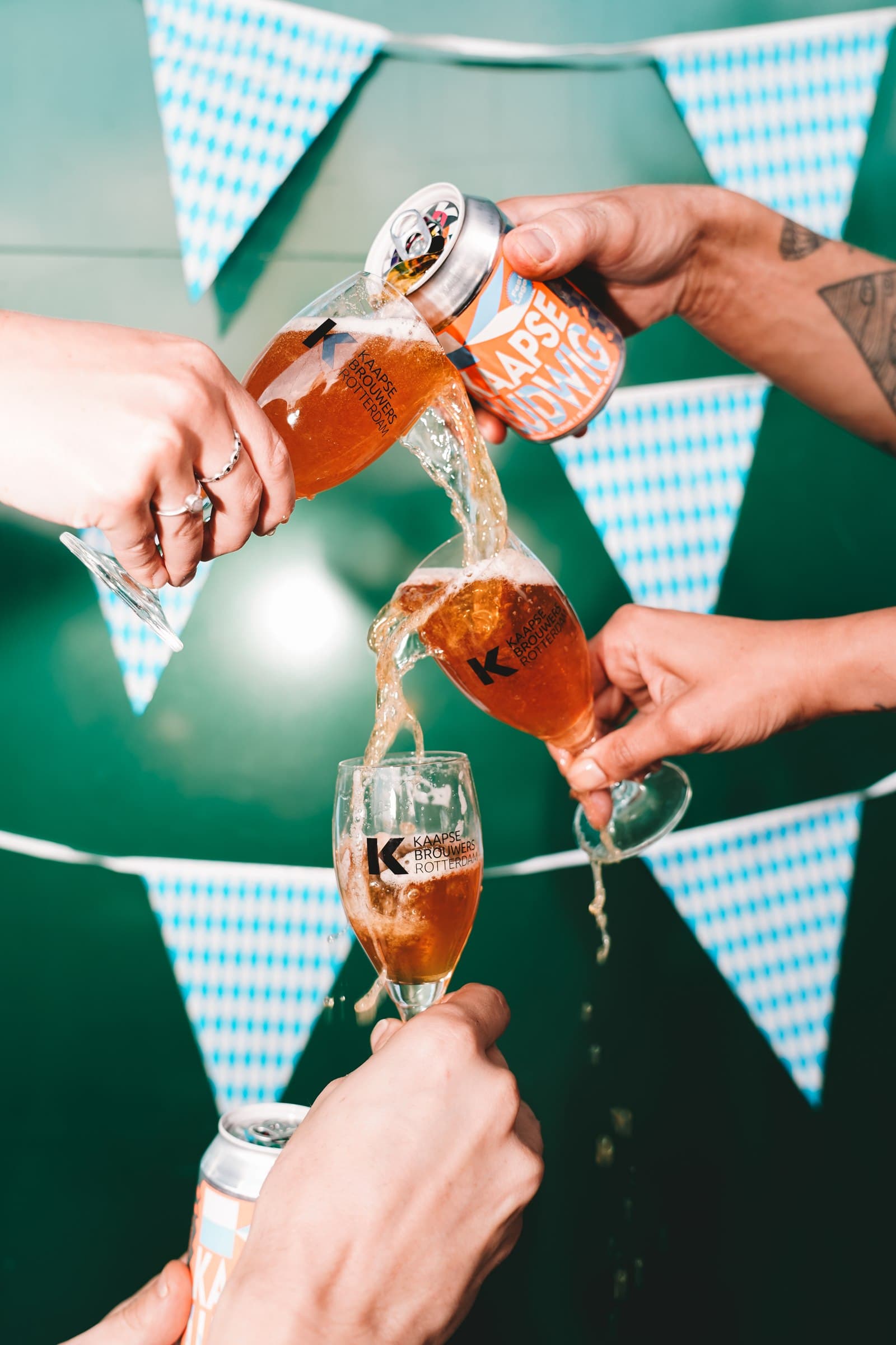 Hands pouring Kaapse beer into a cascade of three glasses against a green backdrop with bunting.