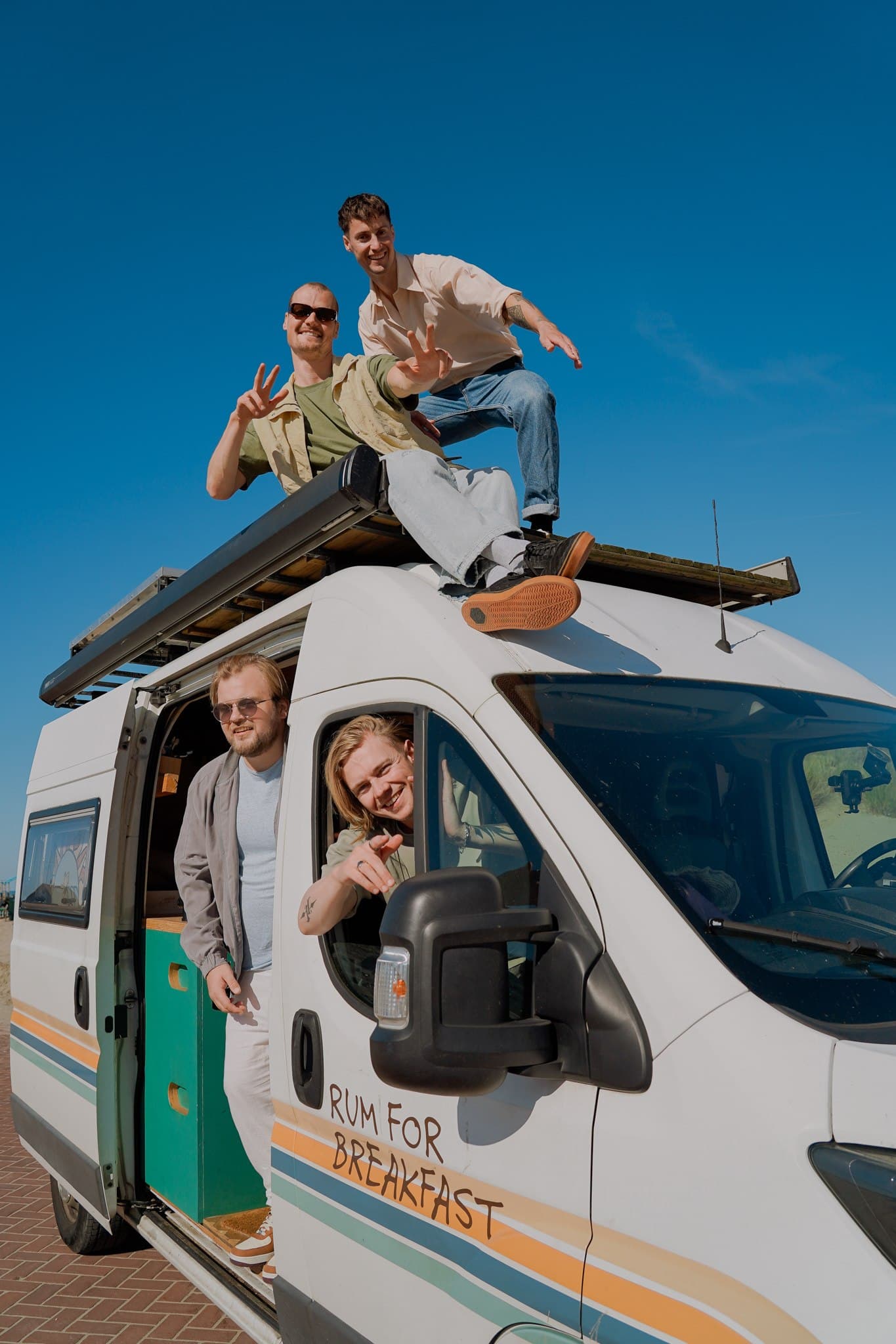 Rum for Breakfast posing in and on their white campervan under a clear blue sky.