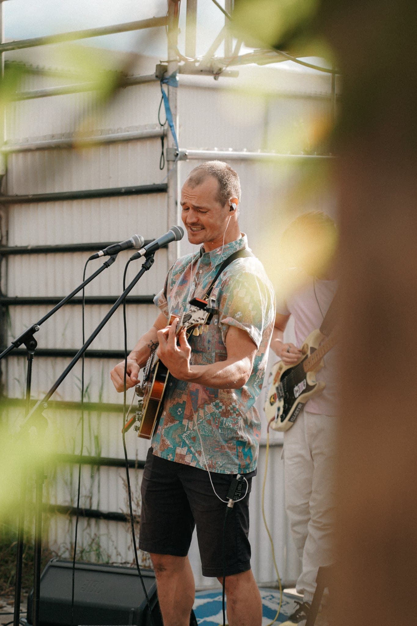A musician performing on an outdoor stage, seen through foreground foliage.