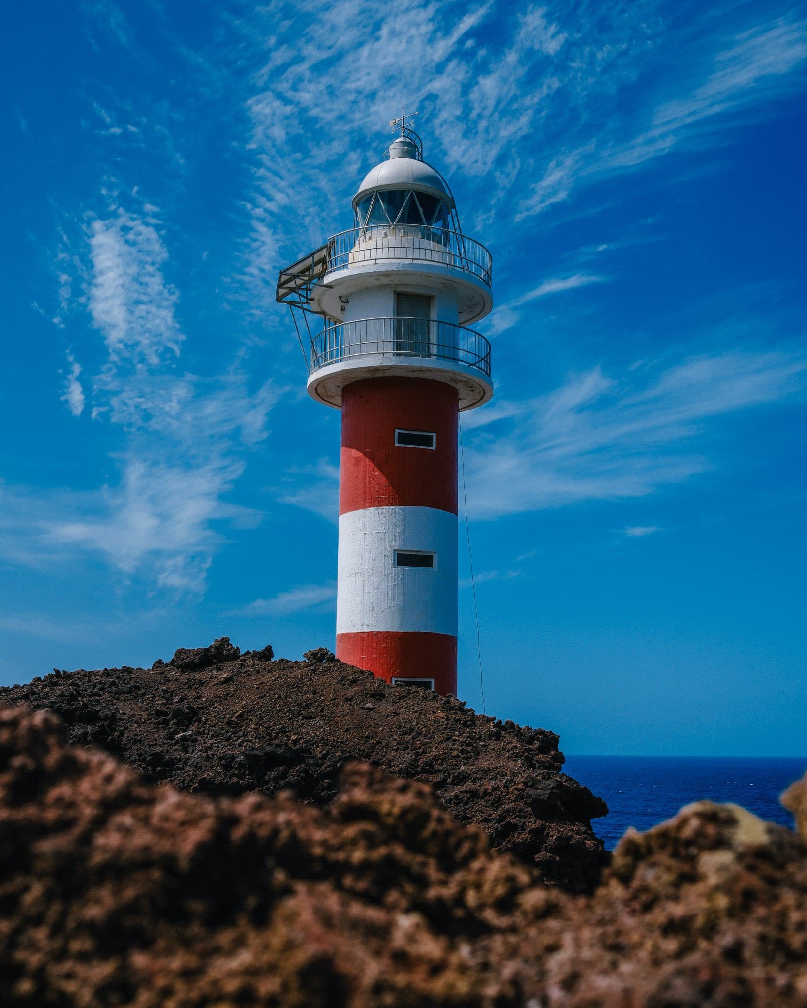 Red-and-white lighthouse above dark volcanic rocks under a blue sky.