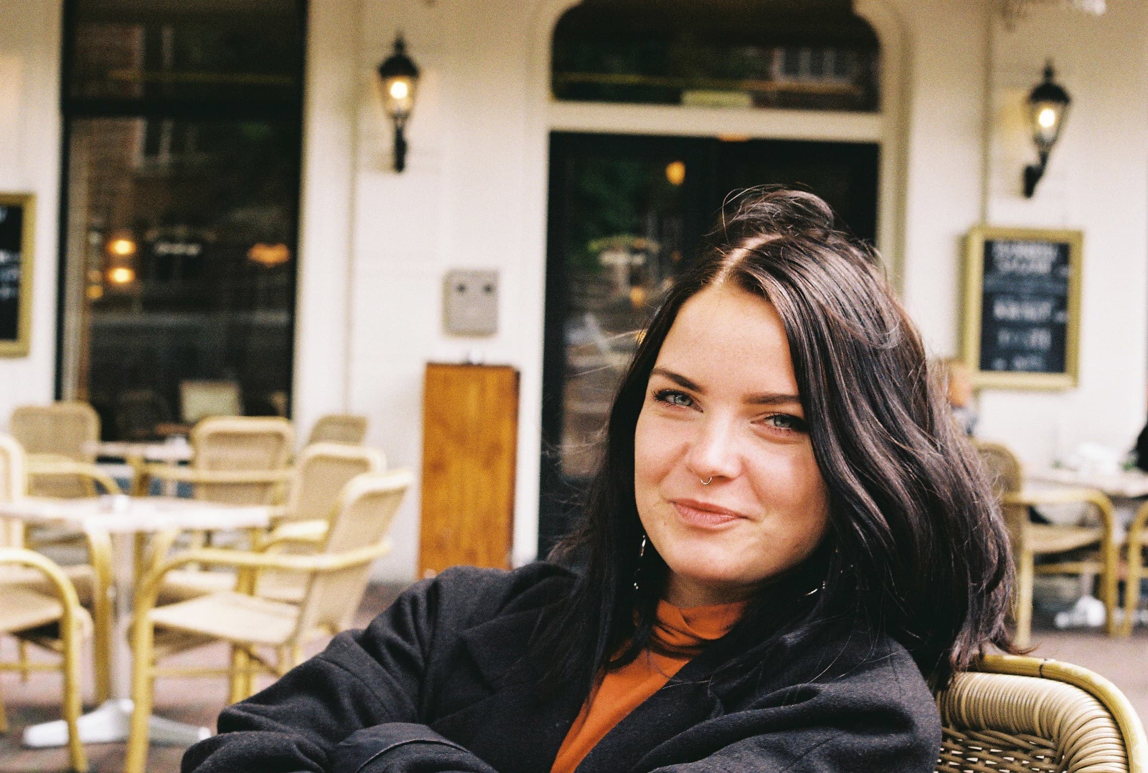 Mara sitting outside at a café, wearing a black jacket and an orange top.